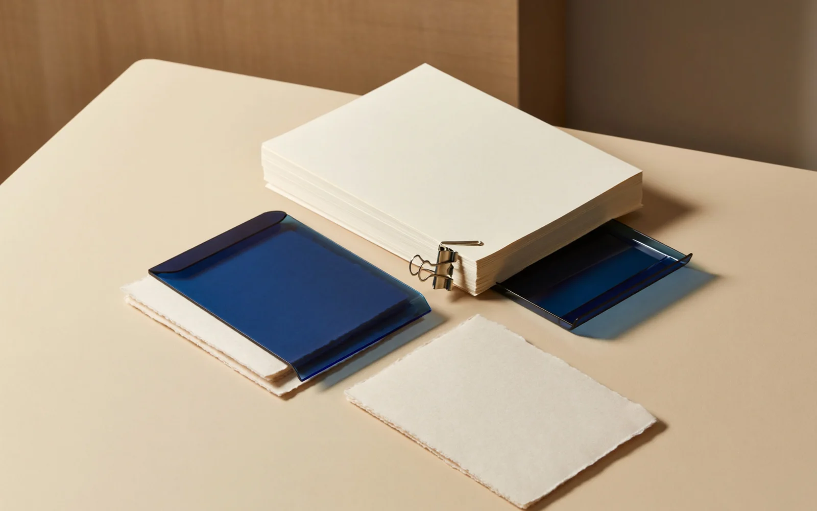 Ivory paper sheets and a steel clip arranged beside cobalt-blue filing trays on a warm tabletop.